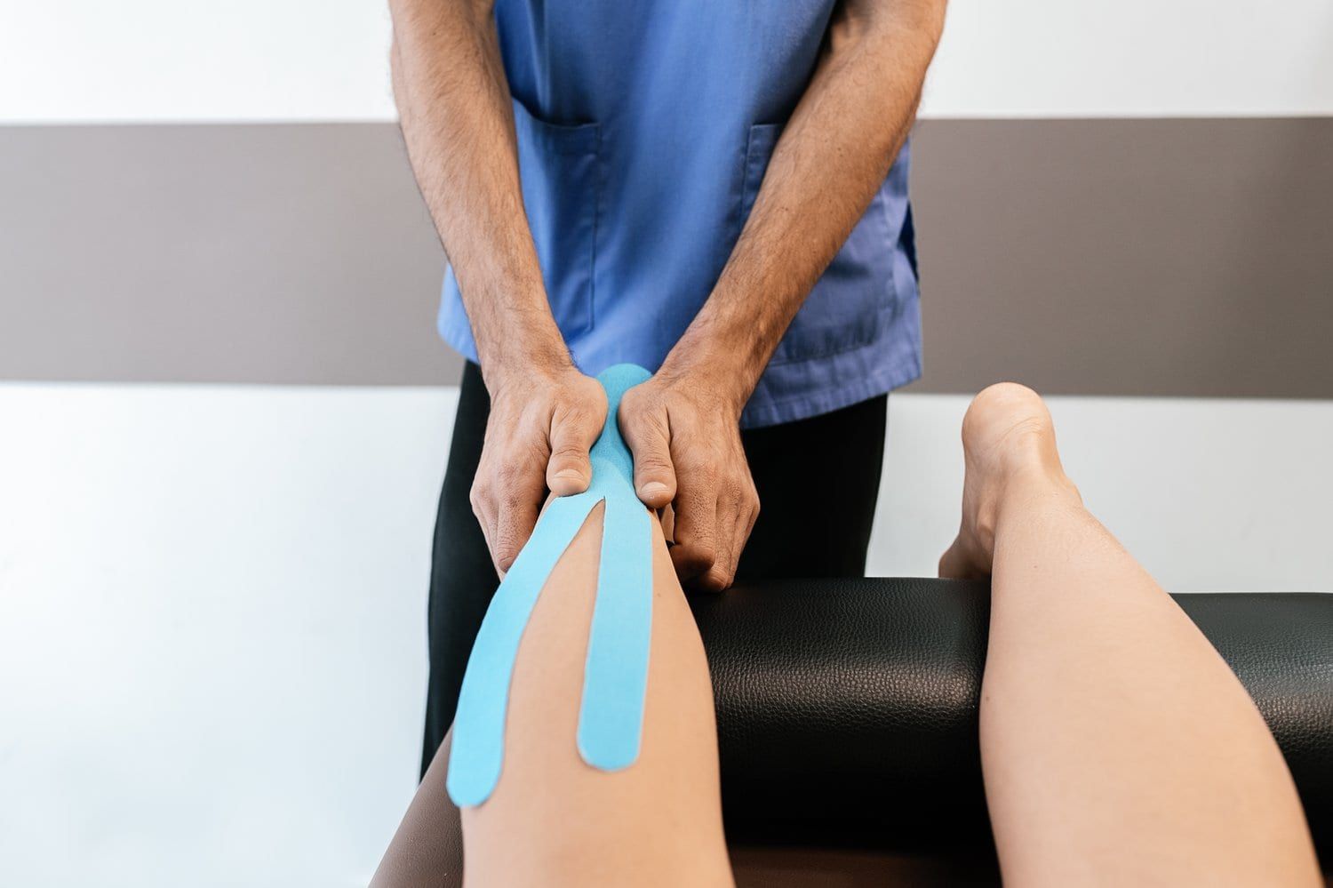 Fizjoterapeuta przykleja niebieską taśmę kinezjologiczną na kolano pacjenta siedzącego na kozetce